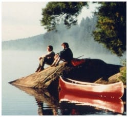 Man and woman sitting on a rock looking over a lake with a canoe in the foreground.
