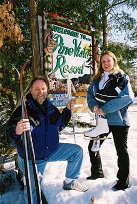 Woman and man posing with Pine Vista Resort sign, ice skates and skis.