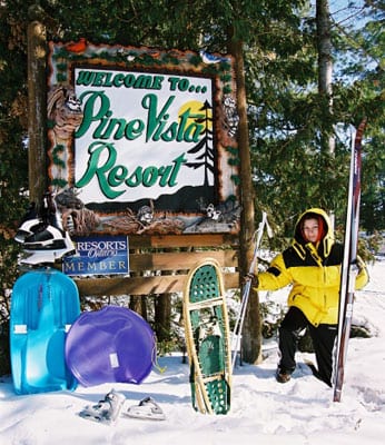 Woman posing with Pine Vista sign, skis, snowshoes, and sleds.