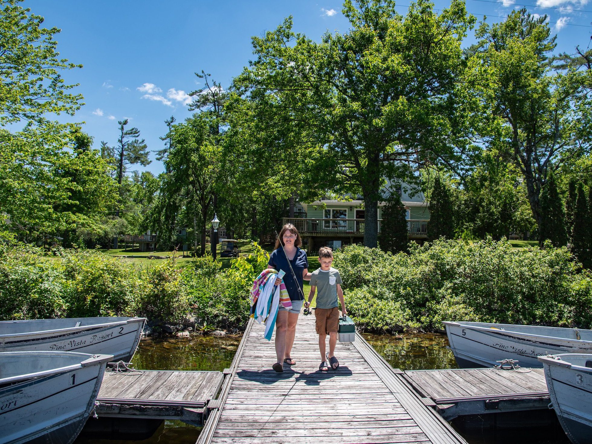 A woman and child walk down a dock, boats on either side and greenery behind them