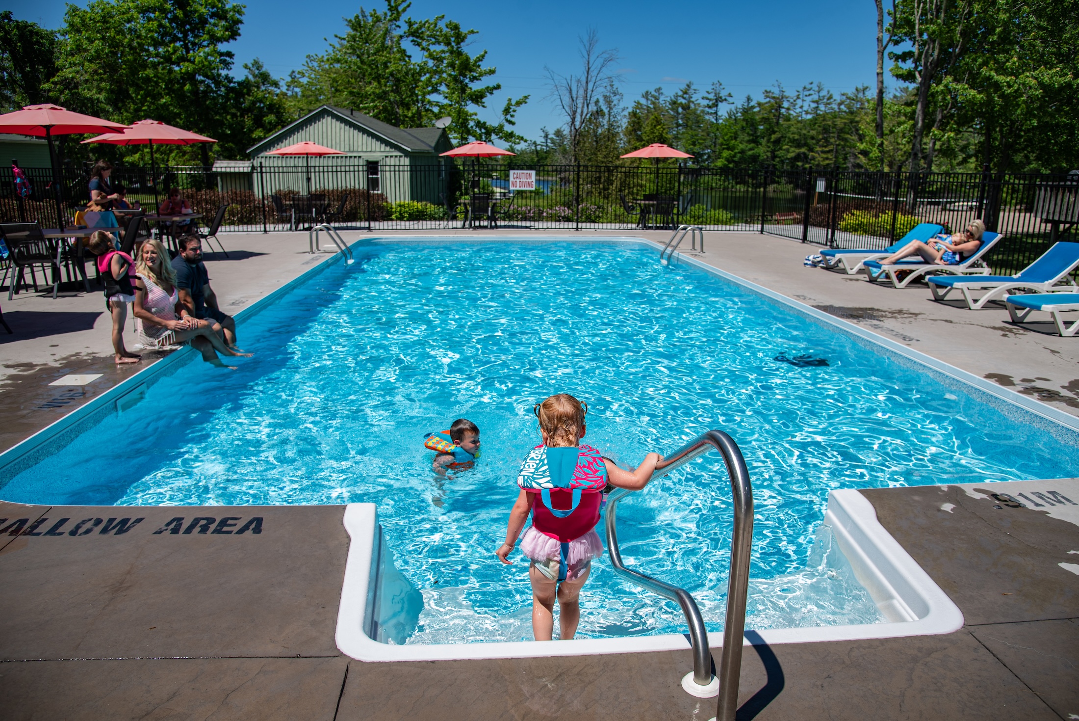 A child enters a blue swimming pool, holding on to the rail, as others look on